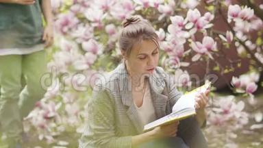 女人在一棵盛开的粉红色玉兰花的灌木背景上看书。 一个卷曲的儿子走到她面前，给了一个蒲公英